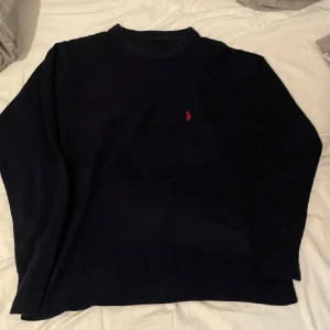 Mörkblå kashmirtröja från Ralph Lauren - Exklusiv mörkblå stickad tröja från Ralph Lauren i 100% kashmir. Klassisk rund halsringning och långärmad modell med den ikoniska röda Polo-loggan broderad på bröstet. Perfekt för dig som gillar stilrena och lyxiga plagg.