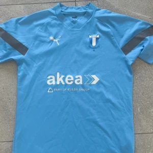 Malmö FF matchtröja Puma ljusblå - Ljusblå Malmö FF fotbollströja från Puma med klubbmärke på bröstet och vit Puma-logga. Tryck med sponsor 'akea' på framsidan. Tröjan har korta ärmar med svarta detaljer och är tillverkad i ett lätt, ventilerande material perfekt för träning eller match.