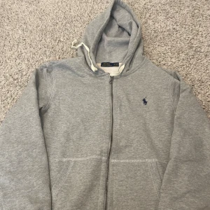 Grå hoodie från Polo Ralph Lauren - Grå hoodie från Polo Ralph Lauren med dragkedja och klassisk blå broderad logga på bröstet. Tröjan har huva med snörning och två fickor framtill. Tillverkad i mjuk bomull, perfekt för chill dagar eller när du vill ha en clean look. Den har aldrig använts och är helt ny. Pris kan diskuteras