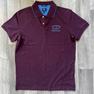 GANT vinröd pikétröja - Snygg vinröd pikétröja från GANT med blå detaljer och broderad text på bröstet. Den är helt ny, oanvänd. Modellen är 167/63 