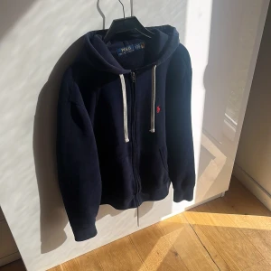 Marinblå hoodie från Polo Ralph Lauren - Säljer en marinblå hoodie från Polo Ralph Lauren med dragkedja och vit snörning i huvan. Mjuk bomullskänsla, perfekt för chill dagar. Kan vara lite dammig, behöver endast avdammas med en klister rulle! Samt lite nopprig men inget som förstör användningen🤩 Nypris 2000 ⬆️ Mvh ItsForfits🌟