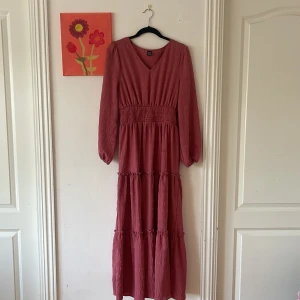 Rosa långklänning för en bekväm sommar  - Långklänning i en snygg dämpad rosa ton från SHEIN. Klänningen har v-ringning, långa puffärmar med resår vid ärmslut och smockad midja. Tyget är lätt strukturerat och kjolen har flera volanger för en flowy look. Perfekt för dig som gillar romantisk stil.