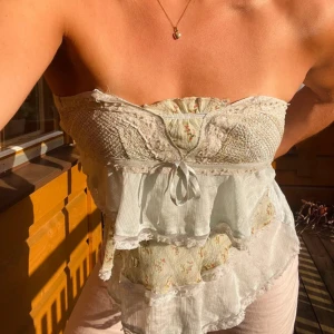 Romantisk blommig bandeau tubtopp - Supersöt bandeau tubtopp i ljusbeige med små blommor och volanger. Toppen har spetsdetaljer, omlottkänsla och en liten rosett framtill. Perfekt för varma dagar när du vill känna dig extra fin.