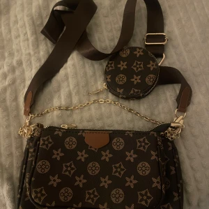 Brun axelväska med monogram och kedja - Snygg brun axelväska med guldiga detaljer och kedja. Väskan har ett monogrammönster i beige och flera fack, inklusive en rund liten väska på axelbandet. Bred justerbar rem i textil och dragkedja upptill. Perfekt för dig som vill ha en trendig och praktisk väska.
