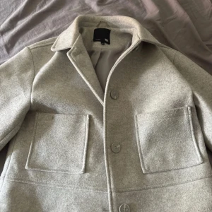 Ljusgrå overshirt med stora fickor - Snygg ljusgrå overshirt från Weekday med klassisk krage, stora framfickor och knäppning framtill. Jackan är i ullblandning och har raka, långa ärmar med knappdetalj vid ärmslutet. Perfekt för lager-på-lager och stilrena outfits.