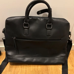 Svart datorväska från Pier One - Snygg svart datorväska från Pier One i stilren design. Väskan är gjord i skinnimitation och har både handtag och avtagbar axelrem. Framsidan har en praktisk dragkedjeficka för småprylar. Perfekt för laptop och viktiga papper.