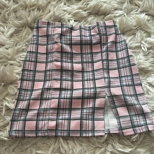 Rutig rosa minikjol med slits -  kortkjol i rosa med svart och vit rutig design. Kjolen har en liten slits framtill som ger en extra snygg detalj. Perfekt för dig som gillar preppy vibes och vill sticka ut lite extra. Passformen är normal
