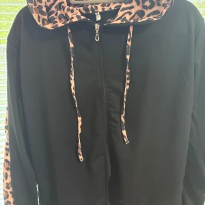 Svart hoodiejacka med leoparddetaljer XL - Svart hoodiejacka med dragkedja och coola leopardmönstrade detaljer på huva, snören och längs ärmarna. Jackan har en avslappnad passform och är perfekt för dig som vill sticka ut lite extra. Materialet känns mjukt och skönt mot huden.
