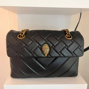 Svart quiltad axelväska från Kurt Geiger - Snygg svart quiltad axelväska från Kurt Geiger med guldfärgad kedja. Väskan är tillverkad i mjukt skinn och har en lyxig känsla med detaljer i guld. Säljer för har bytt till silver. Det är den stora väskan för 3500kr nypris. Kom med bud. Inga skavanker (se bilder på både utsida o insida) kan även skicka med dust bag