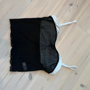 Svart och vit mesh linnetopp - Snygg linnetopp med svart mesh-tyg och vita detaljer. Toppen har tunna, justerbara axelband och en lätt transparent look. Perfekt för lager-på-lager eller att styla med jeans. Enkel men trendig design som sticker ut.