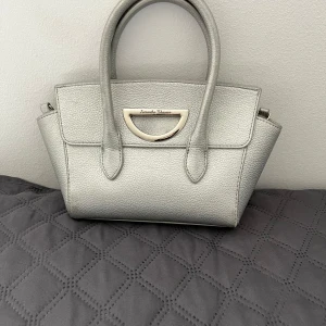 Silver handväska från Samantha Thavasa - Stilren handväska i metallic silver från Samantha Thavasa. Väskan har två handtag, avtagbar axelrem och snyggt metallspänne framtill. Tillverkad i präglat skinn med strukturerad yta och rymligt innerfack. Perfekt för dig som vill ha en trendig och elegant look.