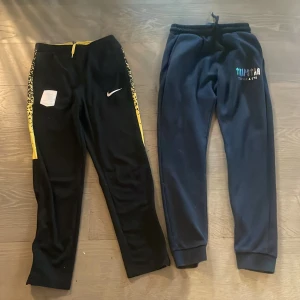 Nike och Trapstar svarta mjukisbyxor - Två par svarta mjukisbyxor, ett från Nike med gula detaljer och snörning samt mönstrade sidor, och ett från Trapstar med tryckt logga i färg. Båda har resår i midjan och är perfekta för chill eller träning. Tryck ej köp nu