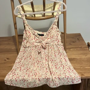 Blommig linnetopp från Jeane Blush - Supersöt linnetopp från Jeane Blush i ljusbeige med små rosa och röda blommor. Toppen har smala axelband, volangdetaljer och en knytning framtill. Luftig och flowy passform, perfekt för varma dagar. Lite genomskinlig 