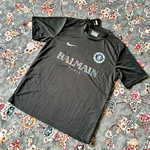 Chelsea x Balmain svart fotbollströja Nike - Svart fotbollströja med Chelsea FC-märke och Balmain Paris-logga i holografisk stil. Tillverkad i lätt och ventilerande material, klassisk rund hals och korta ärmar. Snygg Nike Swoosh på bröstet. Perfekt för dig som vill sticka ut på planen eller gatan.