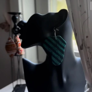 Randigt grönt statementörhänge - Coolt örhänge i en unik, asymmetrisk form med gröna och svarta diagonala ränder. Perfekt för dig som vill sticka ut och addera en edgy touch till din look. Örhänget är stort och hängande, vilket gör det till ett riktigt statement-smycke. Följ oss för mer på Instagram; @shop_with_tyra