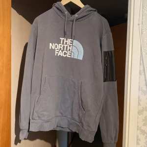 Grå hoodie från The North Face - Säljer en grå hoodie från The North Face med stor vit logga framtill och mindre logga på ryggen. Tröjan har huva med snörning, magficka och en cool svart ficka med dragkedja på ena ärmen. 