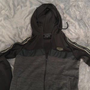 Svart hoodiejacka Supply & Demand M. I nytt skick - Svart hoodiejacka från Supply & Demand med dragkedja, huva och snygga gulddetaljer på ärmarna. Jackan har svarta och mörkgrå partier, samt dekorativa band längs axlarna. Perfekt för en sportig och streetinspirerad stil.
