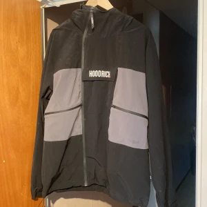 Svart och grå vindjacka från Hoodrich - Säljer en svart och grå vindjacka från Hoodrich i storlek L. Jackan har huva, dragkedja framtill och två stora fickor med snedställda dragkedjor. Stort Hoodrich-tryck på bröstet och meshfoder på insidan. 