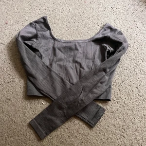 Grå ribbad croppad långärmad topp - Snygg grå croppad topp med ribbad struktur och långa ärmar. Toppen har en bred, rundad urringning och sitter tajt mot kroppen. Perfekt att matcha med höga jeans eller kjol för en stilren look. Aldrig använd