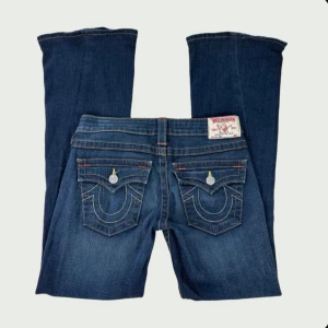 True Religion blå bootcut jeans - Säljer ett par klassiska blå jeans från True Religion med ikoniska bakfickor och kontrastsömmar. Modellen har bootcut-ben och låg midja. Jeansen är i denim med snygga detaljer och logopatch bak. Har två små slitage längst benet.