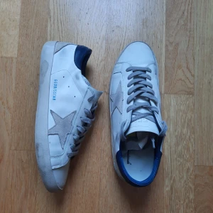 Vita sneakers från Golden Goose - Snygga vita sneakers från Golden Goose med grå mockastjärna på sidan, grå snörning och blå detalj vid hälen. Skorna har platt sula och rund tå, tillverkade i skinn och mocka för en clean men cool look. De är i storlek 42 och är lätt använda och har fortfarande mycket användning kvar i sig! Tyvärr finns inte lådan kvar så de kommer i en annan sko låda.
