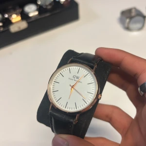 Daniel Wellington svart klocka - Stilren klocka från Daniel Wellington med vit urtavla, svarta markeringar och guldfärgade visare. Klockan har ett klassiskt svart läderarmband och rund boett i guldton. Perfekt accessoar för en clean och tidlös look. OBS batteriet funkar inte!