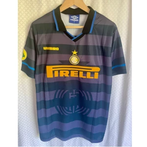Vintage Jersey FC Internazionale Milano  - Klassisk fc Internazionale fotboll hemmatröja säsong 1997/1998 från Umbro med Ronaldo 10 på ryggen. Svart och grå med breda horisontella ränder, gula detaljer och Pirelli-logga på bröstet. Krage med blå och svarta inslag, kortärmad och tillverkad i lätt polyester. Perfekt för samlare eller fans.