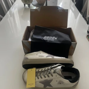 GGDB vita sneakers med grå stjärna - Snygga vita sneakers från Golden Goose med ikonisk grå stjärna på sidan, svarta detaljer vid hälen och grå snörning. Skorna har en platt sula och är tillverkade i skinn med mockadetaljer. Perfekta för dig som gillar en clean men ändå unik look. Dessa skor köpte jag till studenten då de inte passade, inte använda något, storlek 43 men passar även 42. Hör av er vid minsta fundering!!