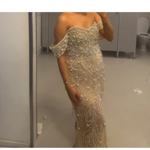 Champagnefärgad pärlklänning offshoulder - Magisk långklänning i champagnefärg med offshoulder och täckt av pärlor och glittriga detaljer. Klänningen har figurnära passform och markerad midja, perfekt för bal eller fest. Materialet är troligtvis polyester eller syntet med lyxig känsla.
