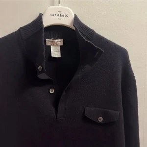 Mörkblå stickad tröja Gran Sasso - Mörkblå stickad tröja från Gran Sasso med ribbad hög krage och knappslå framtill. Tröjan har en bröstficka med lock och knapp samt långa ärmar med ribbade muddar. Perfekt för dig som gillar klassisk och stilren look.