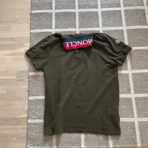 Olivgrön Moncler pikétröja med logga - Snygg olivgrön pikétröja från Moncler med klassisk krage och knappar framtill. Kragen har en cool detalj med Moncler-logga i rött, vitt och blått. Tröjan har korta ärmar och Moncler-märke på ena ärmen. Tillverkad i mjuk bomull. Passar M 