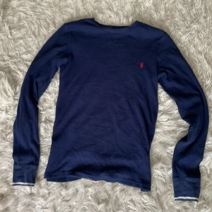 Mörkblå långärmad tröja Polo Ralph Lauren - Mörkblå långärmad tröja från Polo Ralph Lauren med klassisk röd broderad logga på bröstet. Tröjan är stickad och rund i halsen. Tillverkad i mjuk bomull, perfekt för lager-på-lager under höst,vinter och även på sommaren.
