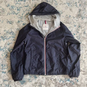 Moncler marinblå vindjacka med huva - Size 164cm Snygg marinblå vindjacka från Moncler med grått innerfoder och huva. Jackan har dragkedja framtill med rödvit detalj, två fickor med knappar och elastiska muddar. Perfekt för dig som vill ha en stilren och fin stil.