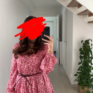 Rosa blommig kortklänning med bälte - Superfin rosa kortklänning med blommigt mönster och v-ringning. Klänningen har långa puffärmar, knappar framtill och ett smalt brunt bälte i midjan. Perfekt för vår och sommar, ger en söt och fräsch look.