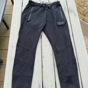 Svarta mjukisbyxor från Woxo 720 - Svarta mjukisbyxor från Woxo 720 med snörning i midjan, dragkedjor på fickorna och en cool patch med text på ena benet. Byxorna har ribbad midja, två bakfickor och är tillverkade i mjukt bomullsmaterial. Perfekta för chill eller träning.