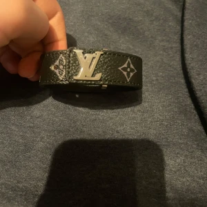 Svart Armband - Snyggt svart armband med klassiskt monogrammönster och stor logga i silver på spännet. Tillverkat i äkta skinn och har detaljer i silverfärgad metall. Perfekt accessoar för att lyfta din outfit.