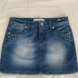 Jeanskjol med detaljer  - Snygg blå jeanskjol med coola nitar på både fram- och bakfickor. Kjolen har klassisk femficksdesign, bälteshällor och knappstängning. Perfekt för dig som gillar en edgy och trendig stil.