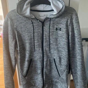 Grå hoodie från Under Armour - Säljer en gråmelerad hoodie från Under Armour med svart logga på bröstet. Tröjan har dragkedja, huva med snörning och två fickor framtill. Perfekt för träning eller chill. Materialet är mjukt och känns sportigt.