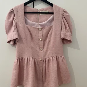 Rosa blus med puffärm och pärlknappar - Supersöt ljusrosa blus från Boohoo med puffiga korta ärmar, volang nertill och pärlknappar framtill. Blusen har små glittriga prickar i tyget och dragkedja i ryggen. Perfekt för dig som gillar romantisk och trendig stil.