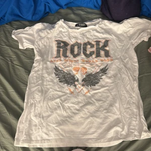 Vit rock t-shirt från Pretty Body - Vit t-shirt från Pretty Body med stort tryck framtill där det står 'ROCK' och coola vingar under. Gjord i 100% bomull, klassisk passform och korta ärmar. 