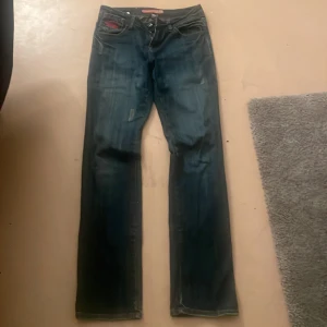 Blå jeans från Reflet med brodyr - Säljer ett par blå jeans från Reflet med raka ben och snyggt röd broderi på bakfickorna. Jeansen har klassisk femficksdesign, normal passform och coola slitningar framtill. Perfekta för dig som gillar detaljer och streetstil.