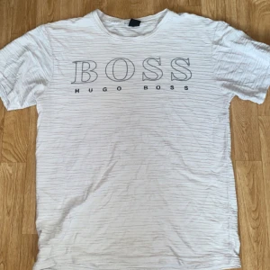 Hugo Boss T-shirt Vit - Vit t-shirt från Hugo Boss med stor BOSS-logga framtill. T-shirten har korta ärmar och rund halsringning. Materialet är bomull och har en lätt strukturerad yta som ger en cool känsla. Perfekt för en clean och stilren look.