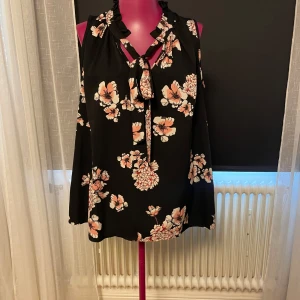 Blommig svart blus med knyt från Lindex - Svart blus från Lindex med blommigt mönster i rosa och beige toner. Blusen har korta ärmar, volangkrage och snygg knytning vid halsen. Luftig och flowy passform. Strl 38