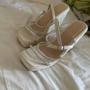 Beiga platåsandaler med strassdetaljer - Snygga sandaler med hög platåklack i flätad jute och remmar dekorerade med glittrande strass. Skorna har fyrkantig tå och justerbar rem runt vristen. Perfekta för sommarens alla tillfällen när du vill sticka ut lite extra.