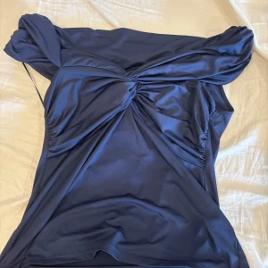 Mörkblå offshoulder topp med knut - Supersnygg mörkblå offshoulder topp med stor knutdetalj framtill. Sitter tight över axlarna och har en glansig finish som ger en lyxig känsla. Perfekt för dig som vill sticka ut med en cool och trendig look.