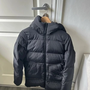 Svart pufferjacka med huva - Svart pufferjacka med huva och dragkedja framtill. Jackan har två stora fickor och en quiltad design som ger en schysst streetkänsla. Perfekt för kalla dagar när du vill hålla värmen och ändå se stilren ut.