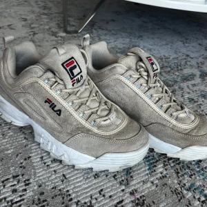 Beige Fila sneakers i mocka - Chunky sneakers från Fila i beige mocka med vit sula och broderad logga i rött, blått och svart på både plös och häl. Skorna har snörning och en robust, sportig design som ger en cool streetstyle-vibe. Storlek 41 men passar som 40.