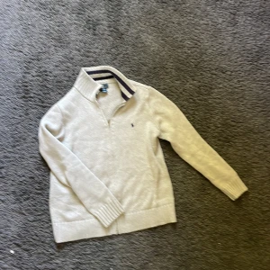 Beige stickad tröja från Ralph Lauren (14-16) - Säljer en beige stickad tröja från Ralph Lauren med halv dragkedja och ribbad krage med mörkblå rand. Tröjan har lång ärm och klassisk passform, perfekt för lager-på-lager. Ikonisk liten logga broderad på bröstet.