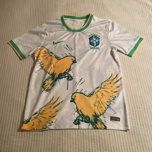 Vit Brasilien fotbollströja Nike - Snygg vit Brasilien fotbollströja från Nike med stora gula fåglar tryckta på framsidan. Tröjan har gröna och gula detaljer vid krage och ärmslut samt det klassiska CBF-emblemet broderat på bröstet. Tillverkad i lätt och ventilerande material, perfekt för match eller träning.