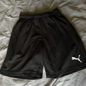 Svarta träningsshorts från Puma - Svarta sportiga shorts från Puma med elastisk midja och klassisk vit logga på ena benet. Tillverkade i lätt och smidigt polyestermaterial som andas, perfekta för träning eller chill. Enkel design utan fickor eller extra detaljer.